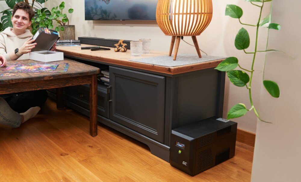 Zasilacz UPS GREENCELL UPSLP700 Czarne urządzenie stoi na podłodze obok szafki pod telewizorem, na której znajduje się lampa, a obok stołu siedzi uśmiechnięty mężczyzna trzymający pudełko z puzzlami, działanie, moc