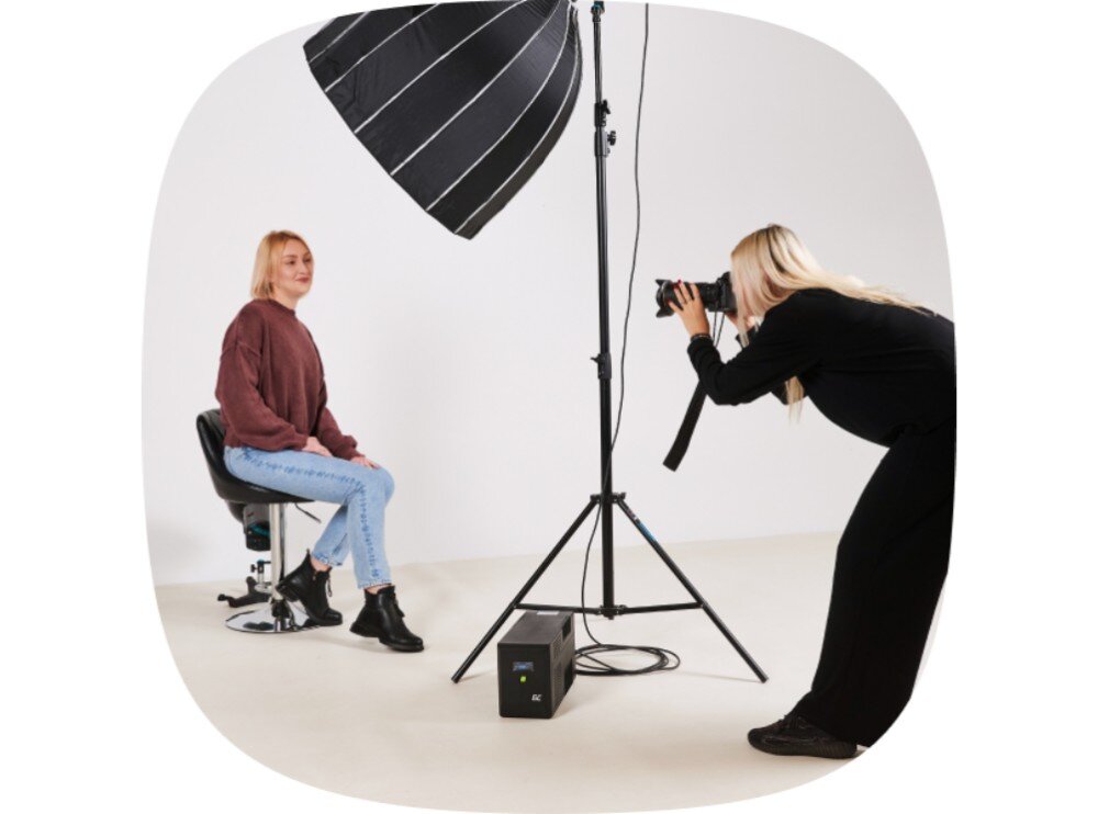 Zasilacz UPS GREENCELL UPSLM900 Kobieta siedzi na stołku w studiu fotograficznym, a druga osoba robi jej zdjęcie aparatem, obok statywu z lampą błyskową stoi czarne urządzenie na podłodze, regulator napięcia, sinusoida
