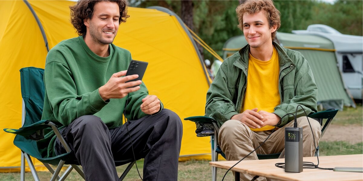 Dwaj uśmiechnięci mężczyźni podczas kempingu, ładujący smartfon za pomocą powerbanku Hama z wyświetlaczem LCD.