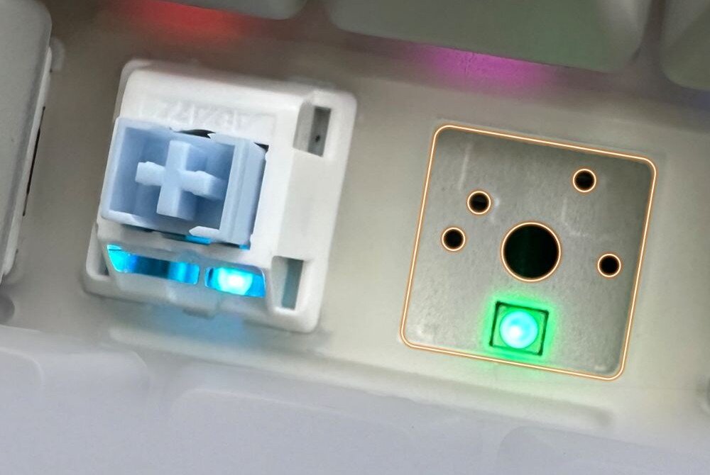 Klawiatura AJAZZ AK820 Pro RGB Zbliżenie na zdemontowany klawisz obok wbudowanej diody LED, ukazujące konstrukcję gniazda i podświetlenie w kolorze niebieskim oraz zielonym, hot swap, wymiana przełączników