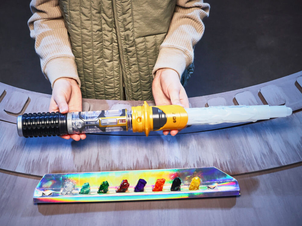 Miecz świetlny HASBRO Star Wars Lightsaber Kyber G07275L0 Zbliżenie na dłoń dziecka naciskającą przycisk na fioletowej obudowie miecza; wewnątrz widoczny jest świecący na niebiesko kryształ kyber.