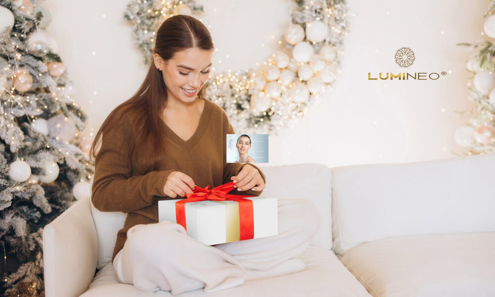 Urządzenie do peelingu kawitacyjnego LUMINEO M22 5w1 + Zestaw nawilżający (Tonik, 2 x Serum, Krem) Uśmiechnięta kobieta w brązowym swetrze i jasnych spodniach siedząca na kanapie w świątecznym otoczeniu, otwierająca białe pudełko przewiązane czerwoną kokardą. Sugeruje to, że zestaw LUMNEO jest idealnym prezentem.