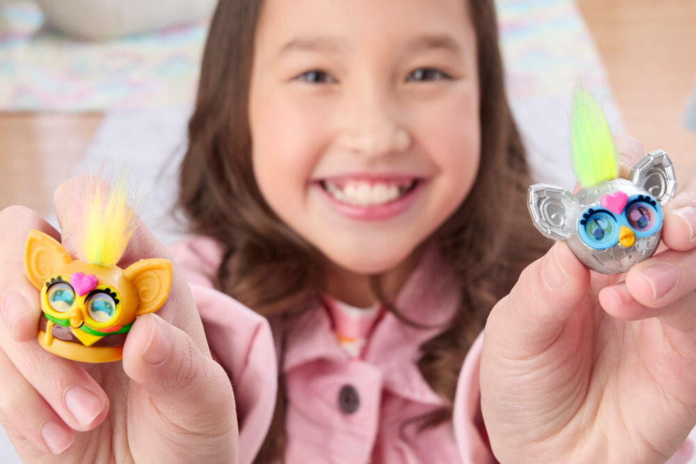 Zestaw figurek HASBRO Furby Minis G19515L0 Uśmiechnięta dziewczynka trzyma w dłoniach dwie figurki Furby Minis z puszystymi czubkami – pomarańczową z żółto-fioletową grzywką i srebrną z zielono-żółtą grzywką.