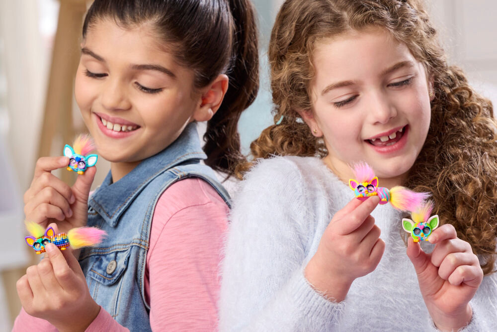 Zestaw figurek HASBRO Furby Minis G19515L0 Dwie roześmiane dziewczynki siedzą tyłem do siebie i trzymają po dwie kolorowe figurki Furby Minis z puszystymi czubkami, oglądając je z bliska.