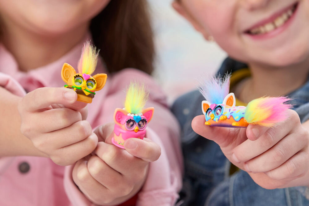 Zestaw figurek HASBRO Furby Minis G19515L0 Zbliżenie na trzy figurki Furby Minis trzymane w dłoniach przez dwie uśmiechnięte dziewczynki – pomarańczową w kształcie burgera, różową w kształcie tortu i kolorową z długim ogonem.