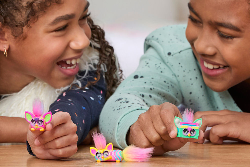 Zestaw figurek HASBRO Furby Minis G19515L0 Dwie roześmiane dziewczynki leżą na brzuchach obok siebie i trzymają po jednej figurce Furby Minis, a trzecia figurka leży na drewnianym stole między nimi.