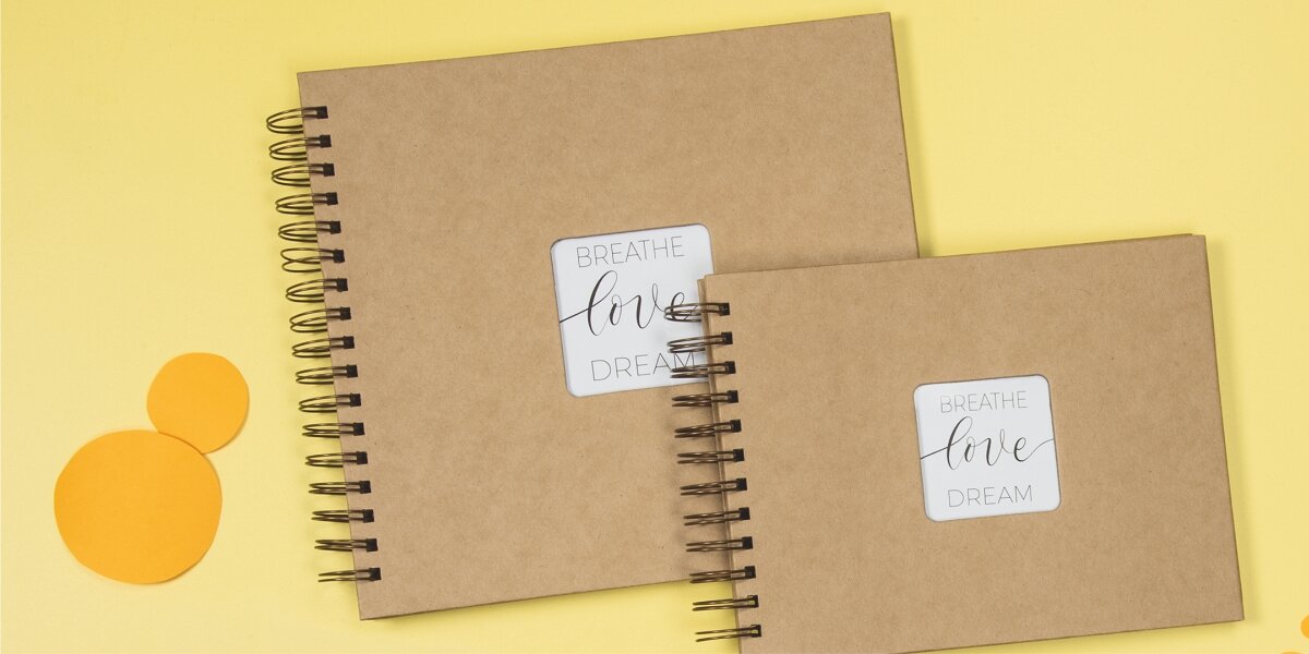 Praktyczna i trwała oprawa Dwa albumy na zdjęcia lub zeszyty ze spiralą w brązowej (kraftowej) oprawie, ułożone jeden na drugim. Na każdej okładce znajduje się biała etykieta z motywującymi słowami: breathe love dream pisanymi ozdobną czcionką.