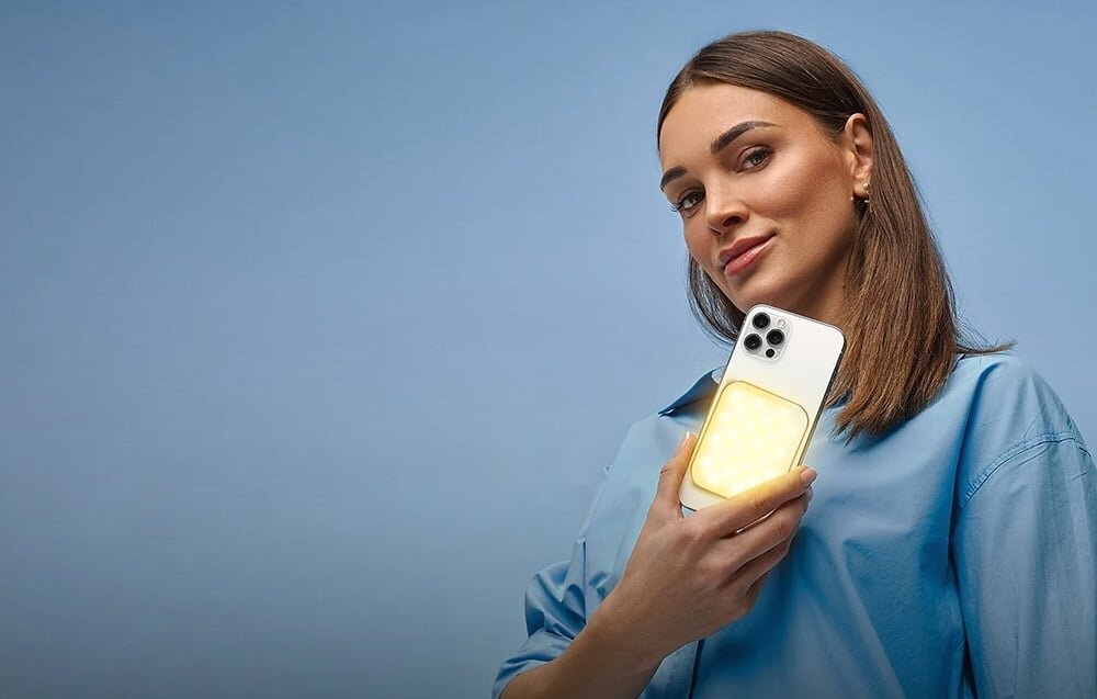 Osoba trzyma smartfon z zamontowaną lampą LED, która emituje jasne światło. Tło jest jednolite, w kolorze niebieskim.