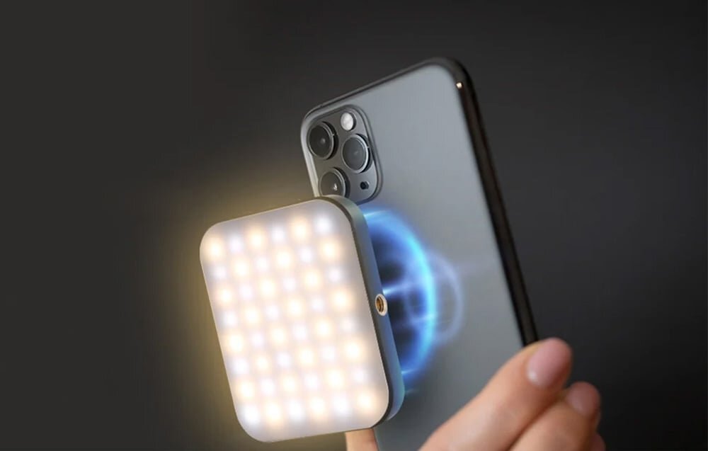Osoba trzyma smartfon z zamontowaną kwadratową lampą LED, która emituje jasne światło. Lampa jest przymocowana magnetycznie do tylnej części telefonu.