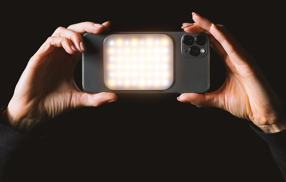 Osoba trzyma smartfon z zamontowaną lampą LED, która emituje jasne światło. Tło jest ciemne, co podkreśla intensywność światła lampy.