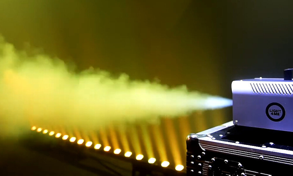 Na obrazie widoczna jest wytwornica dymu z logo \'LIGHT4ME\', emitująca strumień dymu. W tle widać rząd żółtych świateł scenicznych.