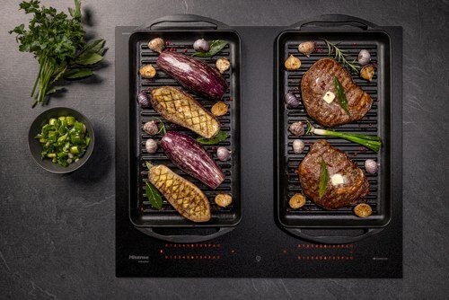 Na płycie indukcyjnej znajdują się dwa grille z warzywami i mięsem. Obok płyty leży miska z pokrojoną zieloną cebulką i pęczek natki pietruszki.