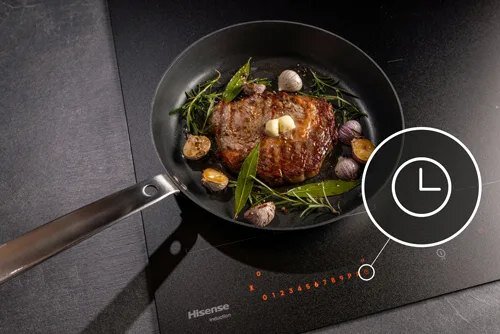 Na płycie indukcyjnej znajduje się patelnia z kawałkiem mięsa, ziołami i czosnkiem. Widoczne jest logo Hisense oraz cyfry od 0 do 9 w kolorze pomarańczowym.