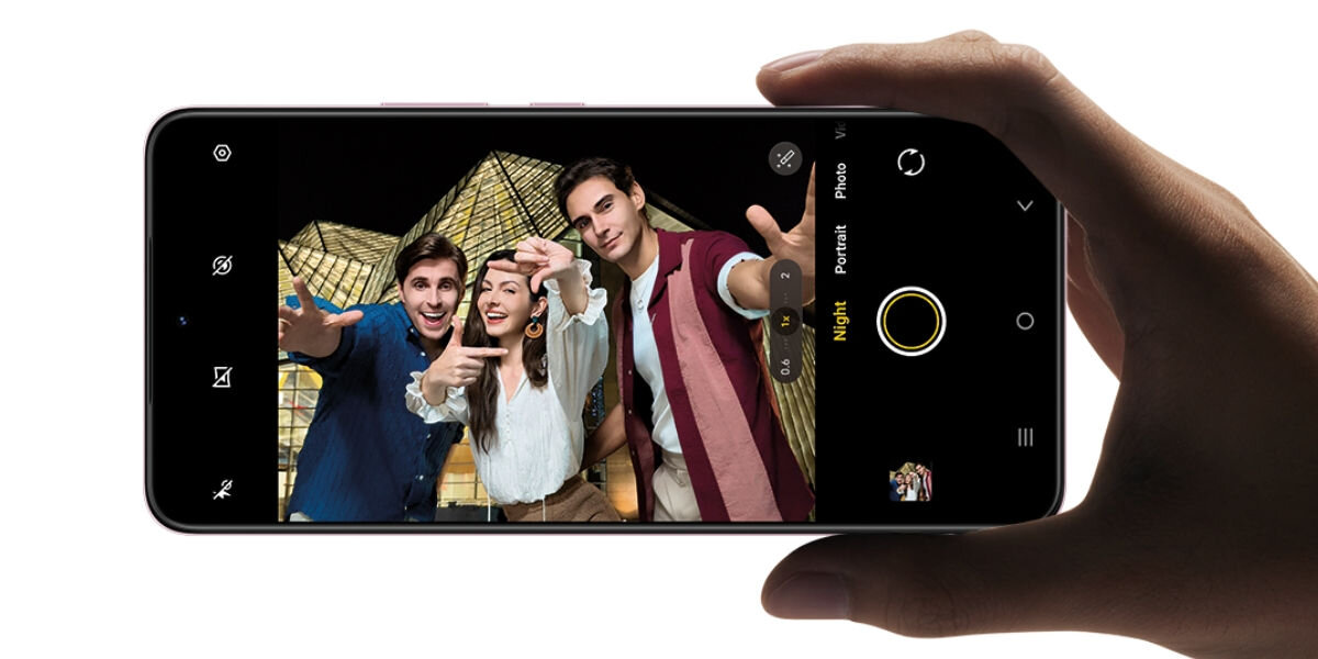 Smartfon VIVO V60 Lite 5G 8/256GB 6.77 120Hz trzymany w jednej dłoni Ujęcie z perspektywy pierwszej osoby, gdzie dłoń trzyma smartfon w poziomie. Na ekranie włączona jest aplikacja aparatu w trybie nocnym (Night). Zdjęcie na ekranie przedstawia troje uśmiechniętych młodych ludzi pozujących na tle podświetlonego, geometrycznego budynku w nocy.