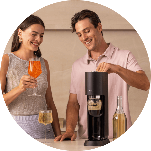 Maszyna SodaStream MIX z mężczyzną nasycającym napój, kobietą trzymającą pomarańczowy koktajl i kieliszkiem wina na blacie