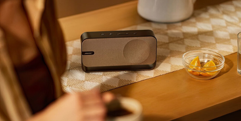 Głośnik mobilny BOSE SoundLink Home - anodyzowane aluminium, maskownica z tkaniny, elegancki design, głośnik mobilny stojący na komodzie w pomieszczeniu, osoba trzymająca filiżankę