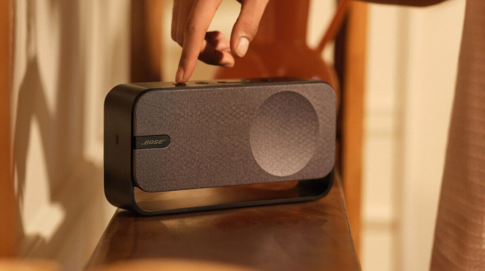 Głośnik mobilny BOSE SoundLink Home - czysty dźwięk, głęboki bas, przetworniki audio wysokiej jakości, głośnik mobilny stojący na półce w pomieszczeniu, ręka trzymająca palec na włączniku
