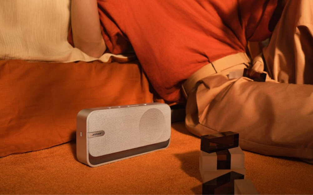 Głośnik mobilny BOSE SoundLink Home - połączenie stereo, dwa głośniki, szerokie brzmienie przestrzenne, głośnik mobilny stojący na podłodze w pomieszczeniu, osoba siedząca obok głośnika