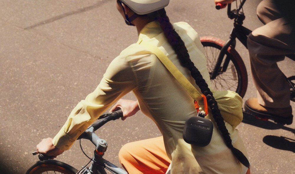 Głośnik mobilny BOSE SoundLink Micro 2nd Gen czysty dźwięk głęboki bas kompaktowy format, osoba jadąca na rowerze z przewieszonym na ramieniu głośnikiem