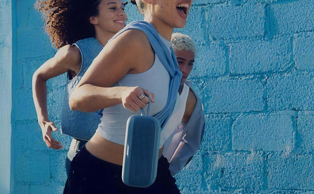 Głośnik mobilny BOSE SoundLink Plus z paskiem do noszenia przenośny głośnik Bluetooth, głośnik stojący na dachu samochodu, osoby biegnące w jednym kierunku, osoba trzymająca głośnik za pasek