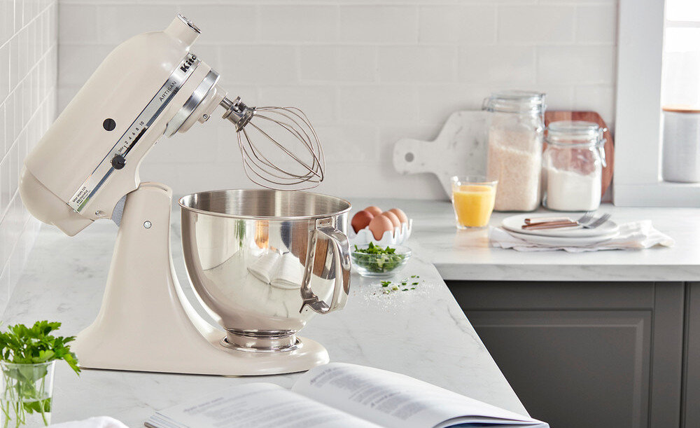 Na blacie kuchennym stoi porcelanowy robot kuchenny planetarny z metalową misą i trzepaczką. W tle widoczne są słoiki z produktami spożywczymi, jajka, szklanka soku i talerze z sztućcami.