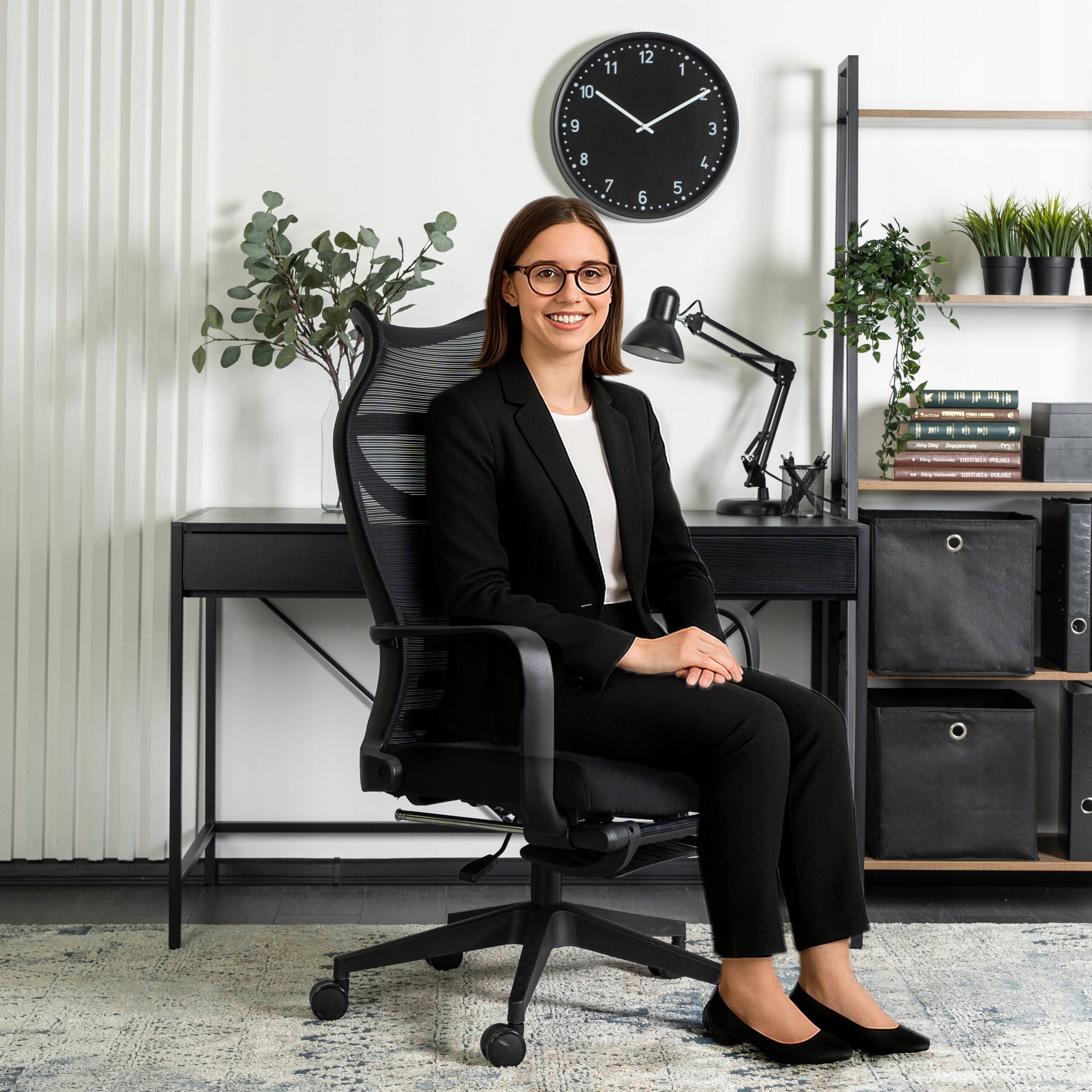 Osoba siedzi na czarnym, ergonomicznym krześle obrotowym przy biurku z lampką i rośliną. Na ścianie wisi zegar wskazujący godzinę 10:10.