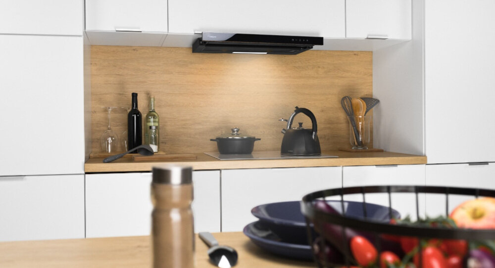 OKAP AKPO WK-7 LIGHT GLASS TOUCH 60 Na zdjęciu widać jasną zabudowę kuchenną z drewnianym blatem i panelem ściennym, nad którymi zamontowano podszafkowy okap o wąskiej, czarnej listwie. Na blacie stoją m.in. butelki z winem, czajnik, garnek oraz pojemnik z przyborami kuchennymi, a na pierwszym planie widoczny jest fragment stołu z przyprawnikiem i miską z warzywami