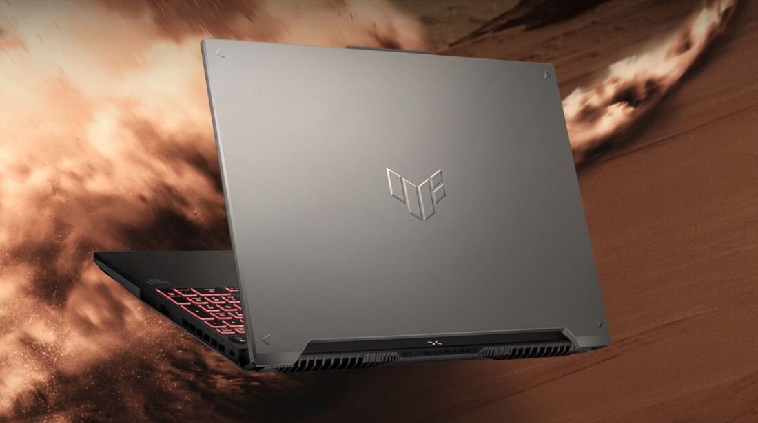 Laptop z metaliczną obudową, na której widoczne jest logo TUF. W tle dynamiczna, piaskowa sceneria.
