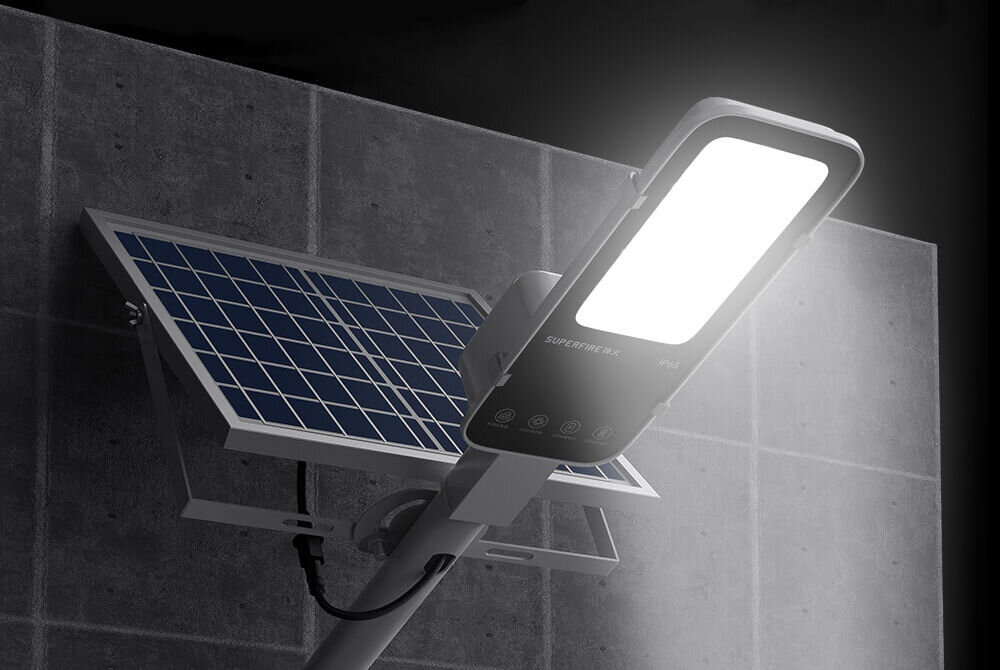 Na obrazie widoczna jest lampa solarna zamontowana na ścianie, z dużym panelem słonecznym i świecącym jasnym światłem. Na lampie znajduje się napis \'SUPERFIRE FF5-E\'.