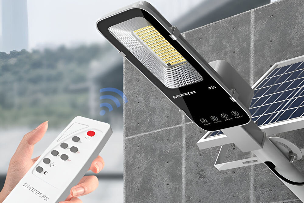 Na obrazie widoczna jest lampa solarna zamontowana na ścianie z panelem słonecznym oraz pilot zdalnego sterowania trzymany w dłoni. Na lampie widoczny jest napis \'SUPERFIRE FF5-E\'.