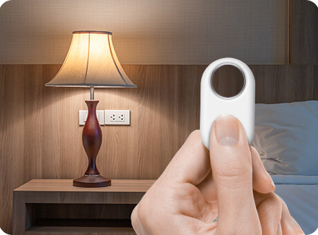 Lokalizator Samsung Galaxy SmartTag2 w dłoni. Osoba trzymająca wciska przycisk na urządzeniu. W tle nowocześnie urządzona sypialnia, w której na szafce nocnej stoi zapalona lampka w klasycznym stylu.