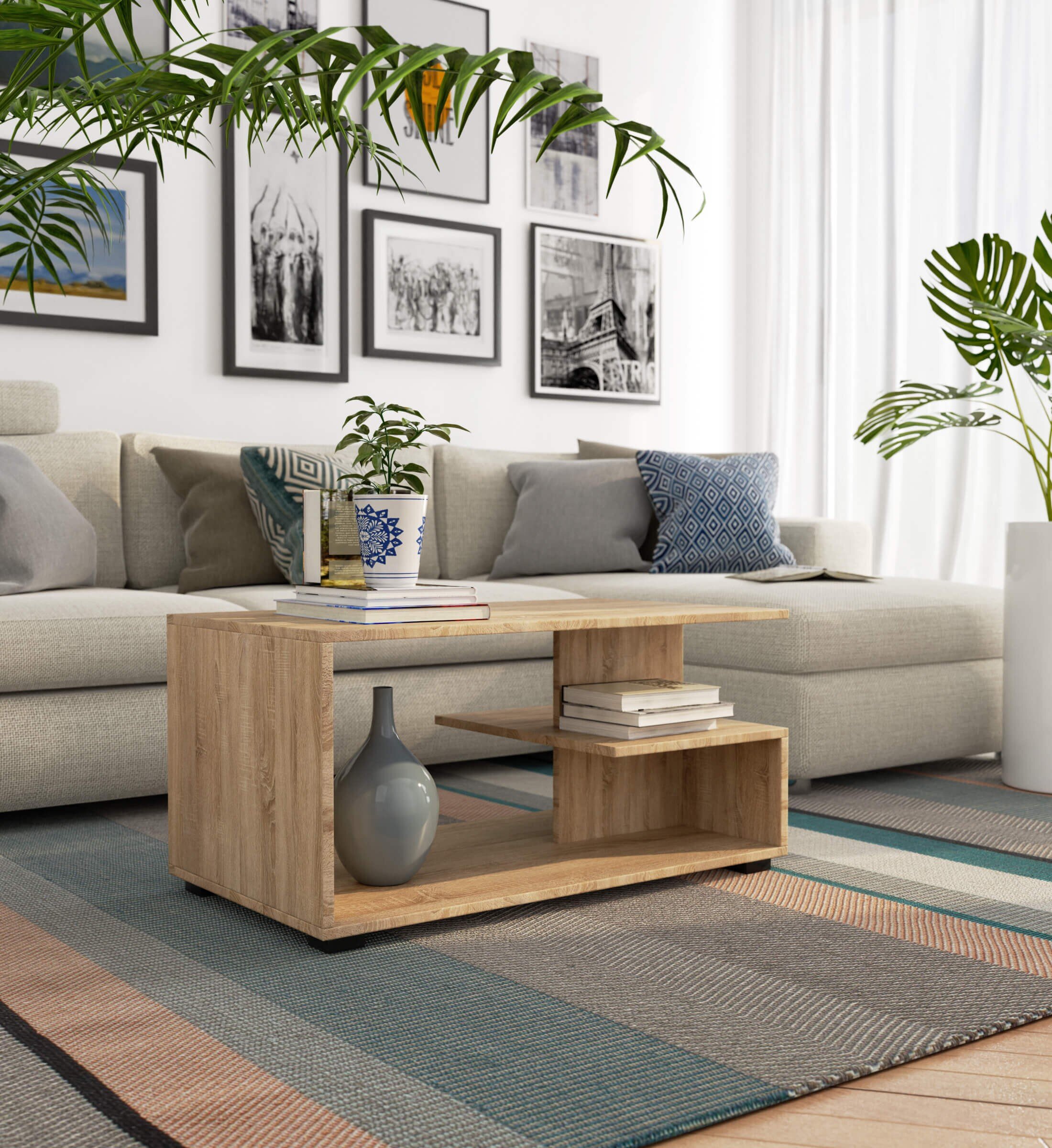 W salonie znajduje się stolik kawowy w kolorze dąb sonoma z matowym wykończeniem, na którym stoi wazon z rośliną oraz kilka książek. W tle widoczna jest jasna sofa z poduszkami oraz ściana z czarno-białymi obrazami.