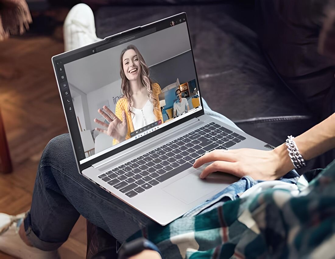 Osoba trzyma laptopa, na ekranie którego widać wideorozmowę z dwiema osobami. Klawiatura laptopa jest widoczna, a osoba korzystająca z urządzenia ma na sobie koszulę w kratę.