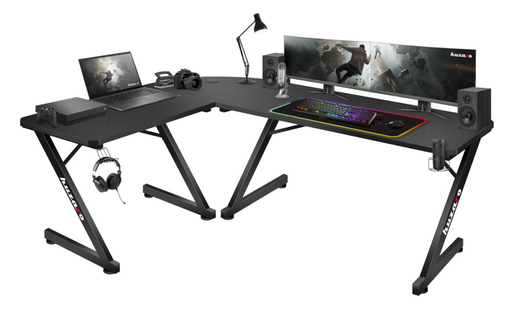 Na obrazie znajduje się czarne biurko gamingowe z dużym monitorem, klawiaturą RGB, lampką, słuchawkami i głośnikami. Na biurku widoczny jest napis \'HUZARO\'.