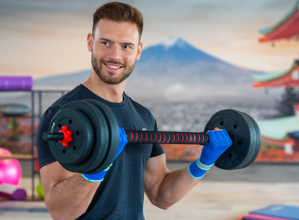 Osoba trzyma zestaw hantli z czarnymi talerzami i czerwonymi elementami na tle krajobrazu z górą. W tle widoczne są elementy wyposażenia siłowni i dekoracje w stylu azjatyckim.