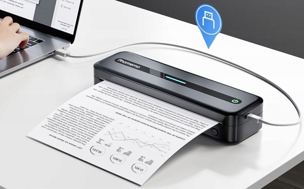 Drukarka PHOMEMO M832 Złącze USB Szary wbudowany akumulator o pojemności 2600 mAh, do 3-4 godzin pracy bez dostępu do źródła zasilania, ładowanie za pomocą dołączonego do zestawu kabla USB-A / USB-C, drukarka na białym blacie podłączona do laptopa, ikona USB-A