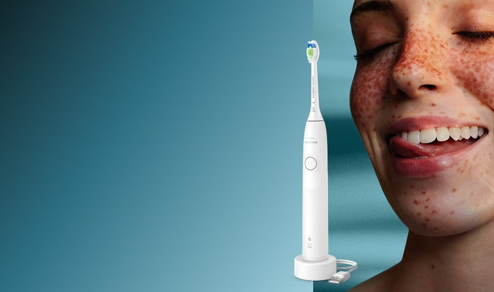 Zbliżenie na twarz uśmiechniętej kobiety, która językiem przesuwa po zębach. Obok szczoteczka soniczna.