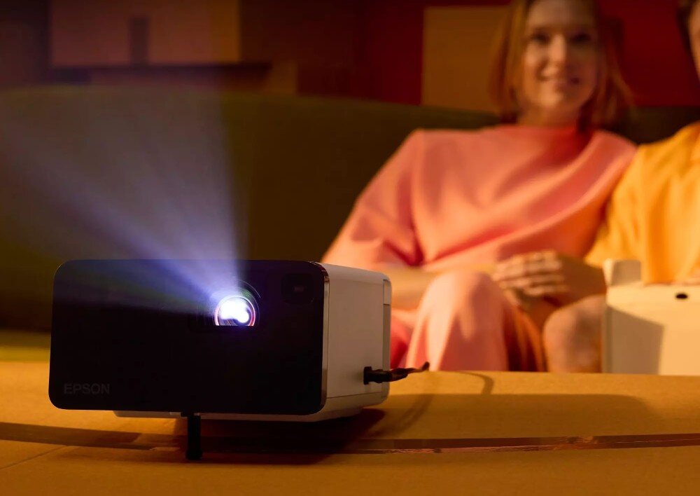 Projektor EPSON EF-21W Projektor w akcji: urządzenie wyświetla obraz w przyciemnionym pomieszczeniu, na pierwszym planie widać soczewkę, z której wydobywa się światło. W tle para siedzi na kanapie, oglądając projekcję, opis
