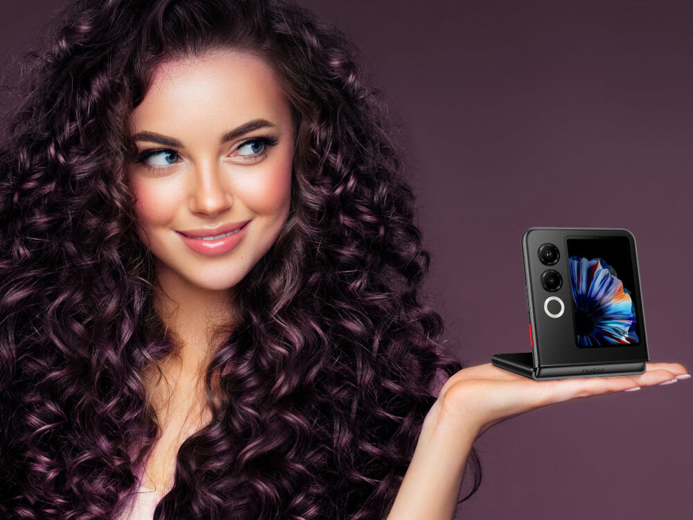 Smartfon NUBIA Flip 2 5G 8/256GB 6.9 120Hz Czarny kompaktowy składany wytrzymały zawias testy wytrzymałości - Kobieta z długimi, ciemnymi, kręconymi włosami, uśmiecha się i patrzy w bok. Na otwartej dłoni, z przodu, trzyma rozłożony smartfon Nubia Flip w kolorze czarnym. Telefon jest złożony pod kątem prostym, z widocznym, małym, zewnętrznym ekranem i dwoma obiektywami aparatu.