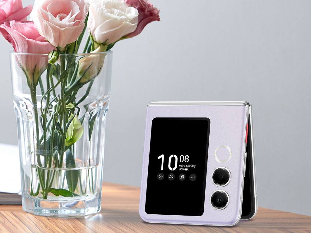 Smartfon NUBIA Flip 2 5G 8/256GB 6.9 120Hz Czarny ekran zewnętrzny OLED 3 cale Always-On Display aplikacje powiadomienia - Złożony smartfon Nubia Flip w kolorze lawendowym, stojący na drewnianym stole obok szklanego wazonu z różowymi i białymi kwiatami (eustomy). Na zewnętrznym ekranie telefonu wyświetlana jest godzina, data i ikony powiadomień.