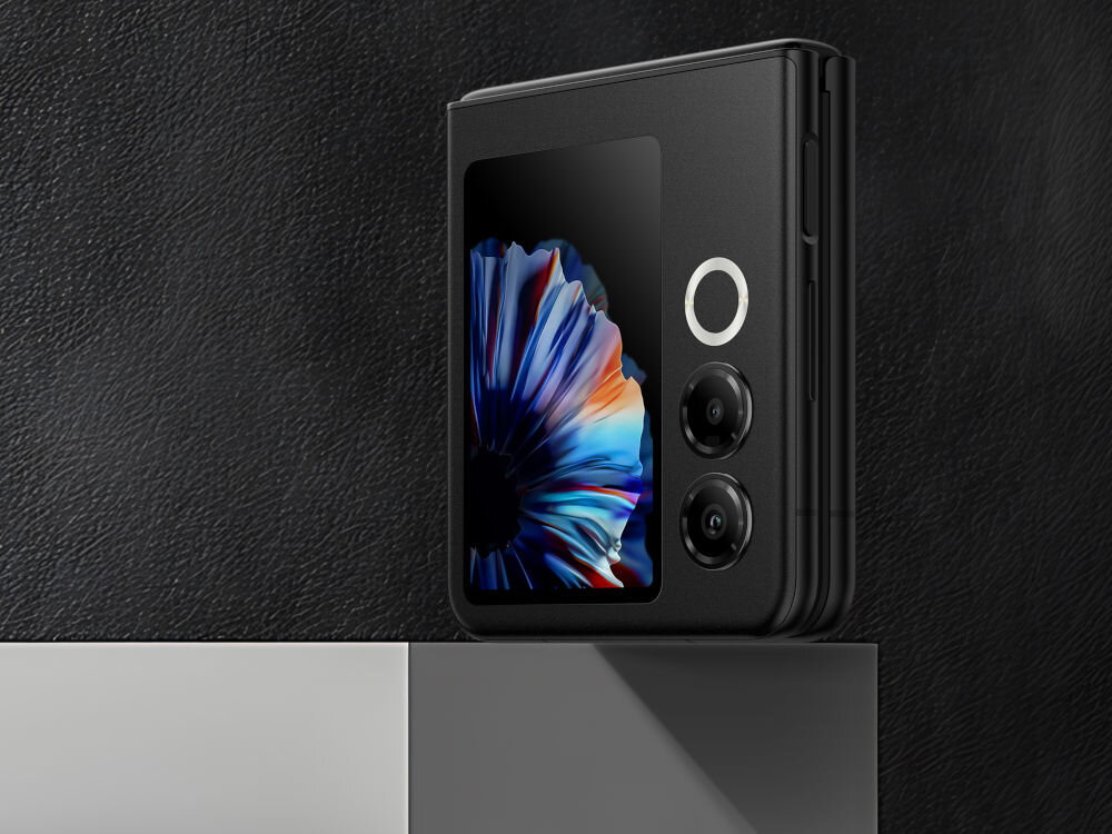 Smartfon NUBIA Flip 2 5G 8/256GB 6.9 120Hz Czarny ekran wewnętrzny OLED 6.9 cala 120Hz 1.5K jasność 1200 nitów ochrona wzroku - zbliżenie na złożony smartfon Nubia Flip w kolorze czarnym, stojący na krawędzi. Widoczny jest kwadratowy zewnętrzny ekran z abstrakcyjną, kolorową tapetą, dwa obiektywy aparatu oraz jasny, okrągły przycisk (prawdopodobnie migawka). Tło jest ciemne i teksturowane (imitacja skóry).
