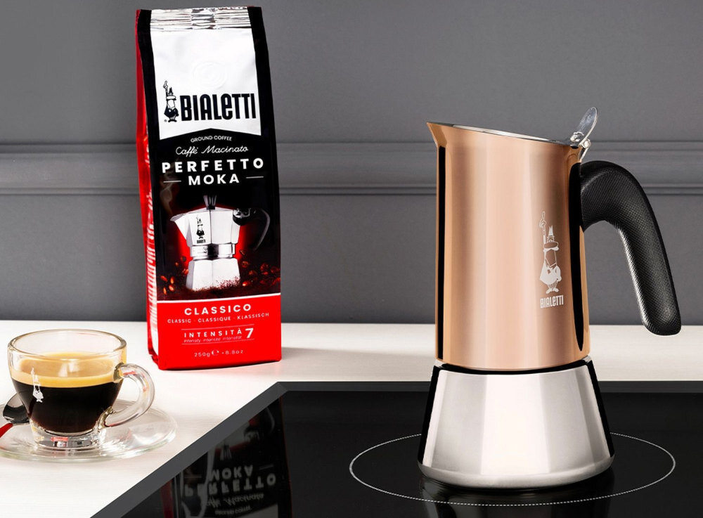 Na obrazie znajduje się miedziana kawiarka z czarnym uchwytem oraz opakowanie kawy z napisem \'BIALETTI Perfetto Moka Classico Intensita 7\'. Obok kawiarki stoi filiżanka z kawą na białym blacie.
