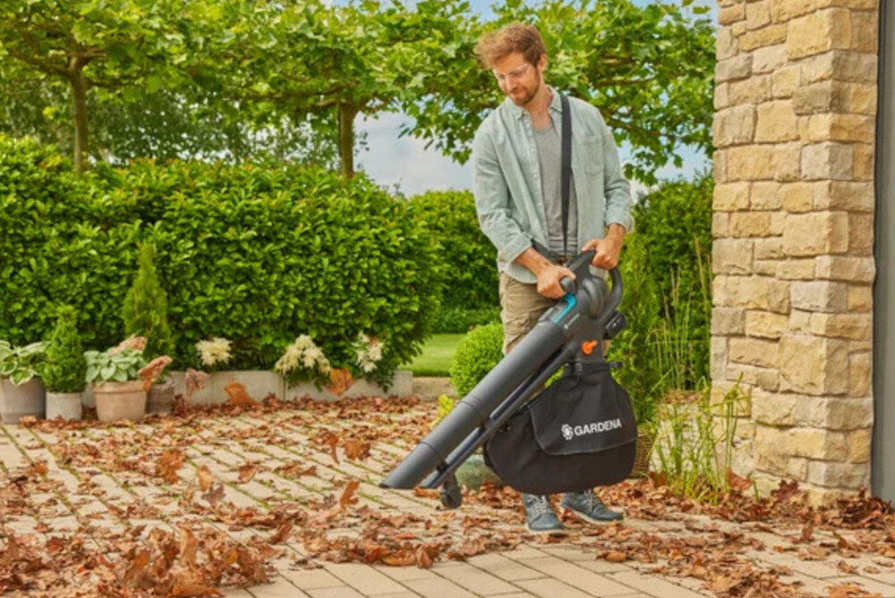 Dmuchawa do liści akumulatorowa GARDENA 18V P4A 14895-55 ogród podjazd osoba odkurza podjazd w tle drzewa krzewy trawnik ułatwia utrzymanie czystości wydajny silnik mocny strumień powietrza porządek wokół domu
