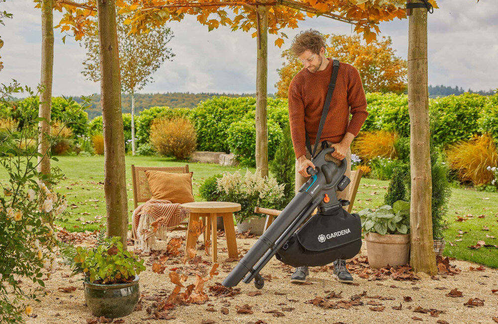 Dmuchawa do liści akumulatorowa GARDENA 18V P4A 14895-20 ogród taras osoba sprząta taras za pomocą dmuchawy w tle rośliny niebo drzewa waga około 4,6 kg wymiary 430 × 405 × 173 mm poziom hałasu wynosi 95 dB(A) wibracje 1,2 m/s² ergonomiczna konstrukcja pasek na ramię