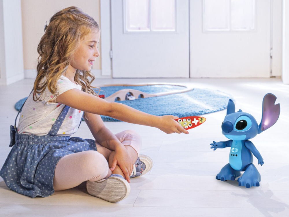 Robot zdalnie sterowany LEXIBOOK Disney Stitch STITCHI17 programowanie sekwencji ruchów quizy edukacyjne mini-opowiadania nauka kodowania zabawka STEM Uśmiechnięta, długowłosa dziewczynka siedzi na podłodze, wyciągając rękę ze zdalnym sterownikiem w kształcie deski surfingowej w kierunku niebieskiego interaktywnego robota-zabawki, przedstawiającego postać Disneya Sticha. W tle widać jasne wnętrze pokoju i niebieski dywanik z zabawką na torach.