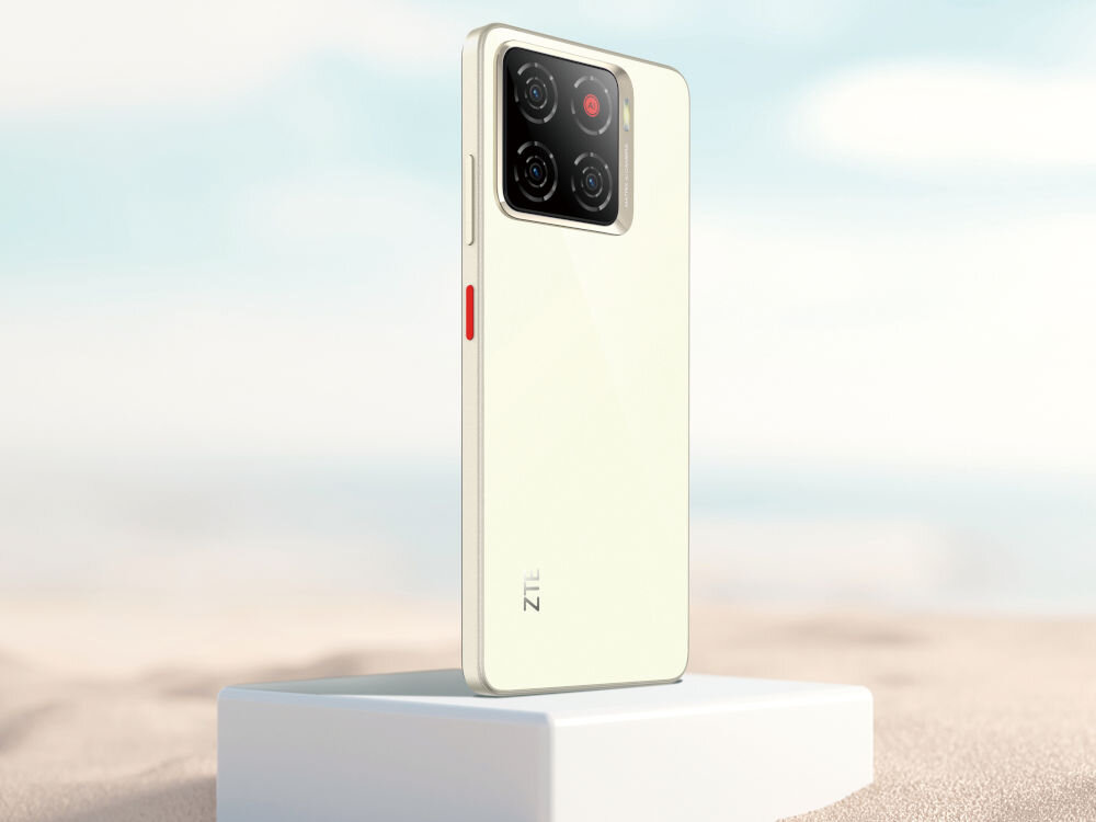 Smartfon ZTE Blade A56 4/128GB 6.75 90Hz Złoty Smartfon w kolorze jasnobeżowym (kości słoniowej) stoi tyłem na białym postumencie na tle nieba i piasku. Widoczny jest kwadratowy moduł z czterema aparatami i czerwonym przyciskiem z boku urządzenia. Z tyłu widnieje logo ZTE.