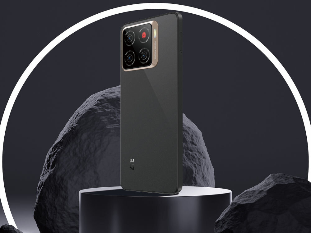 Smartfon ZTE Blade A56 4/128GB 6.75 90Hz Złoty Smartfon w kolorze Złotym z miedzianymi akcentami na module aparatu stoi tyłem na okrągłym, ciemnym postumencie. W tle widoczne są duże, ciemne skały i biały, neonowy okrąg. Z tyłu telefonu widnieje logo ZE (ZTE).