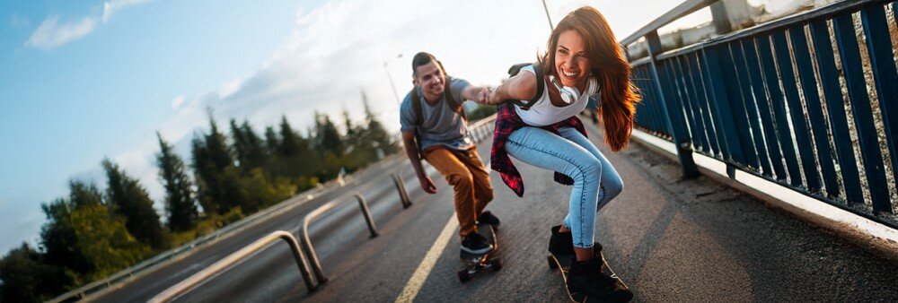 Obrazek przedstawia mężczyznę i kobietę na deskorolkach przejeżdżających przez most