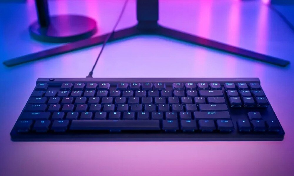 Czarna klawiatura mechaniczna z podświetleniem RGB na biurku, w tle monitor z podstawą w kształcie litery V. Otoczenie jest oświetlone kolorowymi światłami, tworzącymi efektowne tło.