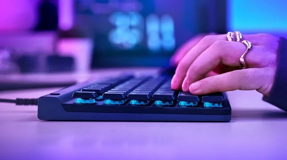 Osoba pisze na czarnej klawiaturze z podświetleniem klawiszy w kolorze niebieskim. W tle widoczne są rozmyte elementy elektroniczne.